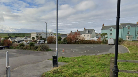 The image shows a mostly empty tarmac car park with just a few vehicles scattered around, bordered by grass, lampposts and a single traffic cone near the marked bays. In the background there are pastel-coloured houses and low buildings, with open countryside stretching out under a cloudy sky.