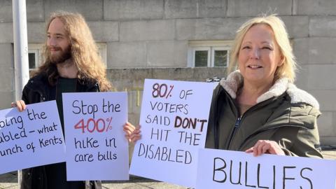 A man with a beard and a blonde lady hold protest signs which read "stop the 400% hike in care bills" and "80& of voters said don't hit the disabled"