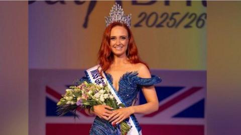 Alice Cutler wearing a dark blue sparkly dress, a sash with the words: "Miss Great Britain" and holding a bunch of flowers and a crown in her hair. She has red hair and is stood on stage after winning.