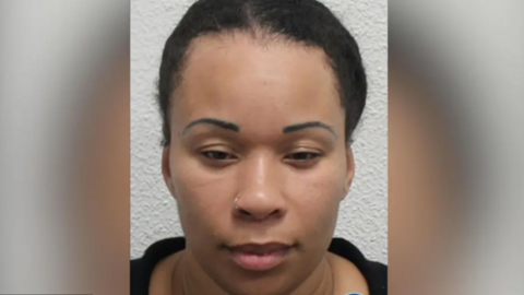 A woman with pulled-back black curly hair looking at the camera in a police mugshot. She is wearing a black top and small gold nose ring.