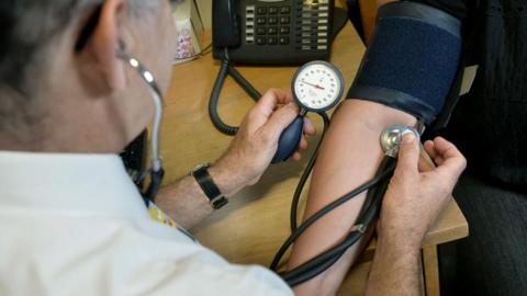 A doctor takes the blood pressure of a patient and listens to their pulse with a stethoscope. A black telephone stands on the desk in the background.