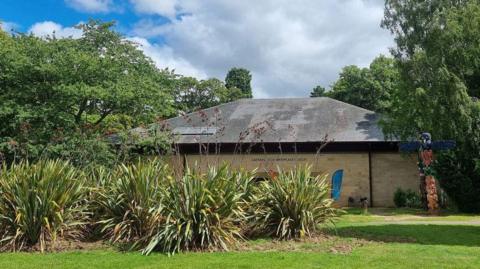 Small one storey building in background with green grass surrounding it. Big bushes in front.