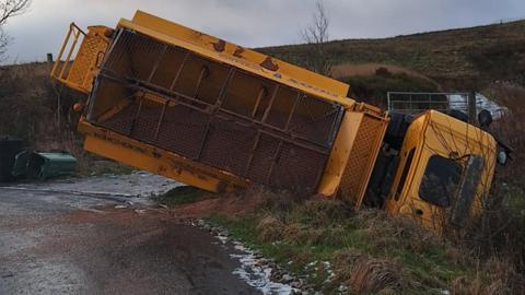 An overturned gritting lorry stuck in a ditch off a road on great, icy, wet moorland countryside