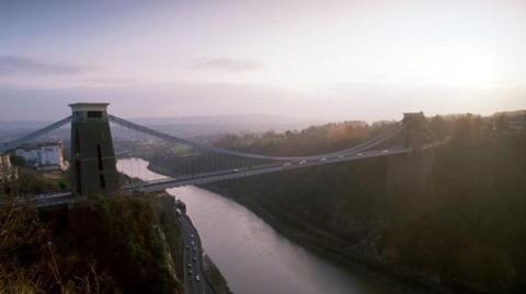 A panoramic view of the Clifton Suspension Bridge in the early morning light. The Bridge has two huge towers at each end. The River Avon is in visible in the gorge below.