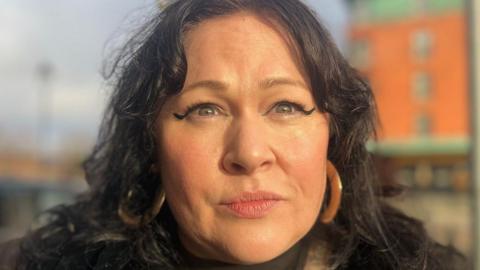 Sara Morrison has long dark hair parted in the middle and is wearing large hoop ear-rings