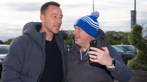 John Terry, wearing a black top and navy hooded coat, poses for a selfie with a Colchester fan outside their ground. The fan is wearing a blue and white-striped bobble hat and a coat.