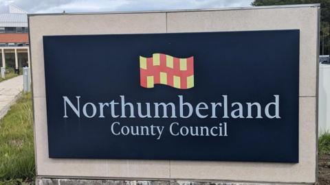 A black sign with white writing which reads 'Northumberland County Council' which sits underneath a red and yellow flag. 