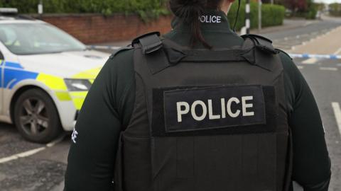 A police officer in a bottle green uniform with a black vest on with 'police' written across it in white writing stands in the middle of the road. A police car sits parked to their left.
