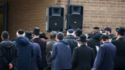 Traditionally dressed mourners at the service, pictured outside with two large speakers relaying the ceremony