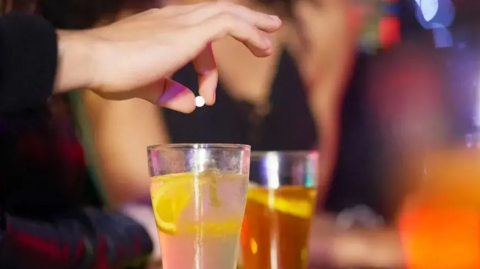 A close-up of an unidentified pill being dropped into a drink. The background is out of focus, but the picture has been taken in a pub or a club.