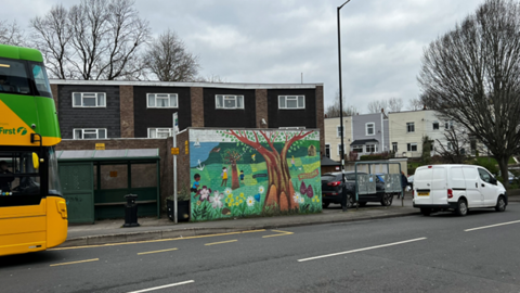 A view of a bus stop on a grey day: red brick building behind with windows and a flat roof. The bus stops have a bright mural painted next to them, of a cheery green park with flowers and children playing. On the left, the front of a yellow and green bus starts to come into view