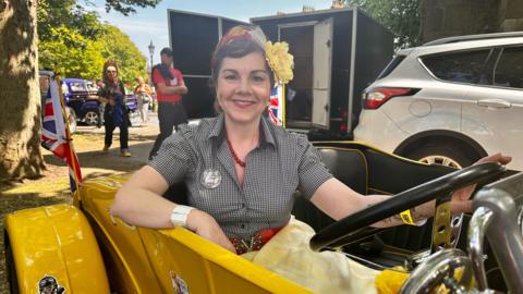 A woman with dark hair, a headscarf and large yellow flower smiles as she sits in black and yellow car. She is wearing a black check dress with a red necklace and red belt.