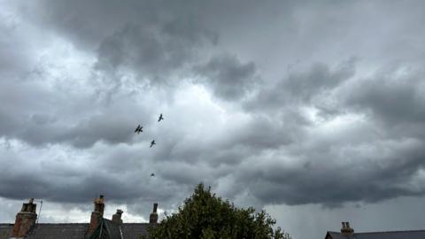Dark grey rain clouds building over a row of houses, five large birds flying amidst the cloud