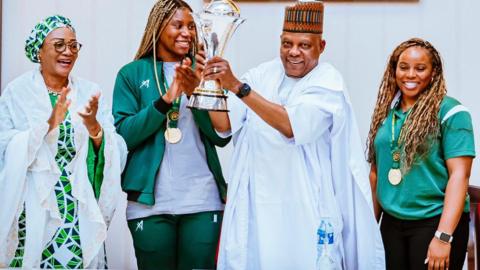 Nigerian Vice-President Kashim Shettima (2nd right) dressed in a white kaftan lifts a trophy with two members of the victorious national female football team either side of him who wear green uniforms and medals. Another female official dressed in the national colours claps to the left of the image.
