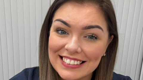 Clare has long dark hair and is smiling at the camera. She has blue eyes and is wearing pink lipstick. She is standing in front of white blinds.