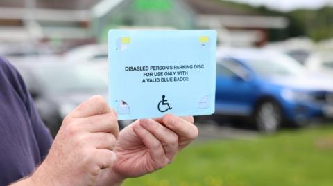 A pair of hands holding a disabled person's blue parking disc, with an out of focus blue car behind it