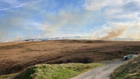 A line of fire and smoke stretching across brown moorland with a police van in white, blue and florescent yellow livery parked at a road junction