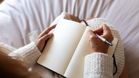 POV high angle view of Unrecognizable young woman wearing a robe sitting on bed writing on journal in cozy bedroom. - stock photo
