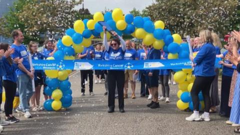 A woman carries a large blue baton towards a thick ribbon which reads "Yorkshire Cancer Research". She is under a balloon arch made up of blue and yellow balloons. A large group of people wearing blue t-shirts are standing around the balloon arch.