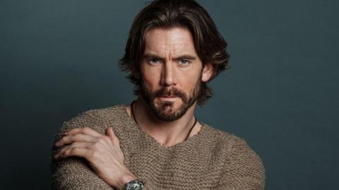 A headshot of James Wilkinson in a brown knitted jumper. He is posing while sat down in front of a dark green background. He has long brown hair and a dark beard.
