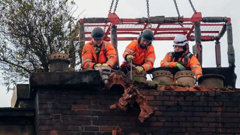 Workers in orange uniforms hack an a brick chimney and scatter bricks. They are on a platform suspended in the air