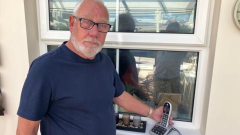 Jim Crawford standing in front of a window in his house with his cordless phone on the windowsill. He has short white hair and a beard and wears glasses. He is looking at the camera with a serious expression.