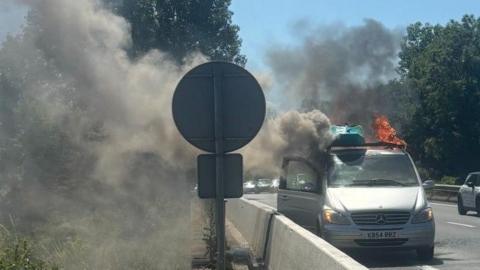 A silver van by the side of a dual carriageway with a kayak on the roof, flames and smoke billowing from it.