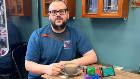 Tim Brogan is sitting at a table with his hand on the handle of a cup of tea, which is on a saucer on the table. He has glasses and a beard and is wearing a blue T-shirt. He is sitting by a teal wall with two wooden glass cabinets behind his head. On the table are colourful dice and small cards.