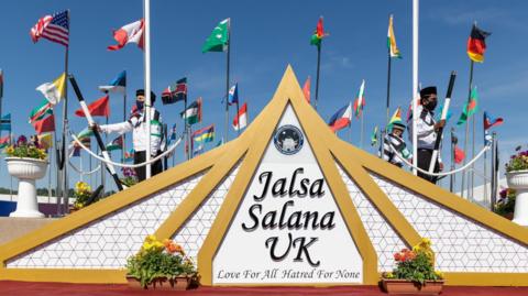 Three men in black and white uniforms pose holding staffs near a sign which reads "Jalsa Salana UK - Love for all, hatred for none". In the background are poles bearing flags from different nations