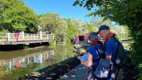 A man and a woman look at a piece of paper while they stand on a canal path on a sunny summer's day. On the canal a boat is travelling towards the camera.