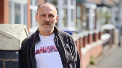 Andy Polshaw, a man wearing a white t-shirt and a black fleece jacket is standing on a street with terraced houses behind him.