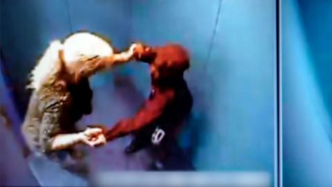 A still from CCTV footage showing a woman with blonde hair in a ponytail grabbing the wrists of a boy wearing a red hoodie as she pushes him into a windowless room with blue padding on all of the walls