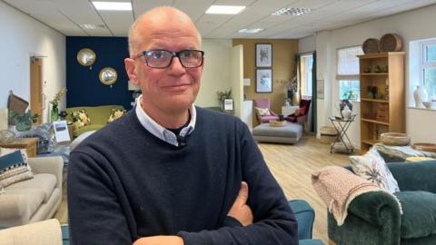 Simon Robinson stands in front of his sofa showroom. He has thick-rimmed black glasses, and wears a blue striped shirt and a blue jumper. In the background, a variety of sofas and staged living room set-ups can be seen.