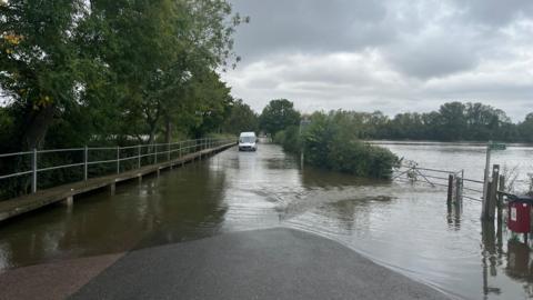 Felmersham bridge is submerged in water. It is hard to distinguish from a large body of water nearby divided by a line of bushes. a van is stuck on the bridge