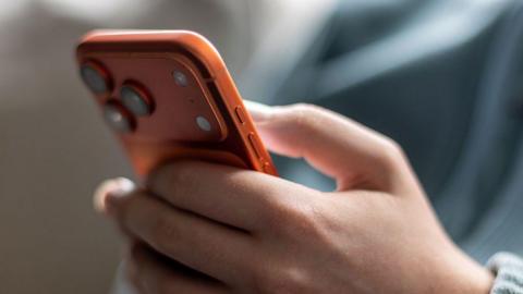 A teenage boy holds an orange iPhone.