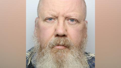 A man with a bald head and long grey beard. He is wearing a blue checked shirt and standing against a grey background