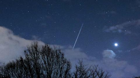 A long streak from a shooting star in a dark, clear night sky with some cloud in the foreground