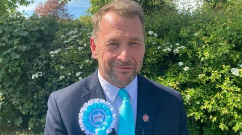 Stephen Atkinson standing outside in front of greenery smiling  in the sunshine. He has short fair hair and stubble and is wearing a blue suite and Reform UK rosette