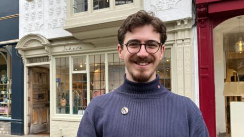Bob Hewis standing on St Mary's Passage, Cambridge. He has waving dark brown hair and a trimmed beard and full moustache. He is wearing black framed glasses and a navy roll-neck jumper.