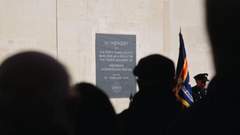 Silhouttes of people stood looking at a grey memorial plaque, against a cream stone wall, that says 'In Memory of the forty three people who died as a result of the tragic accident at Moorgate Underground Station on the 28th February 1975'.