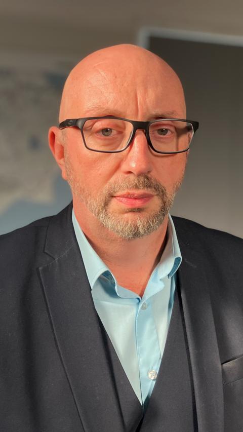 A bald man wearing glasses, a blue shirt and a black jacket looking straight at the camera