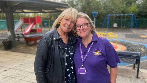 Linda Robson, who is wearing a black leather jacket and a black-and-white blouse, with her arm around Heather Hill, who is wearing a navy blue "Happy Hill"-branded polo shirt. They are both smiling and standing in the Send centre's playground.