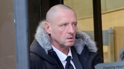 Mark Byrne is pictured looking off to one side in front of a court house. He is bald and clean shaven and wearing a dark suit and tie with a white shirt. He has a black parka on top with a fur-lined hood.
