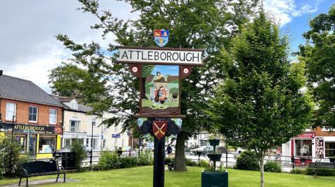 Attleborough town sign. It's on a green in front of green trees, with shops visible in the background.