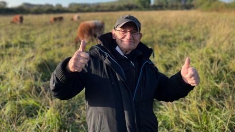 Colin Attenborough is standing in a field holding his thumbs up. He is wearing a dark peaked cap and black coat.