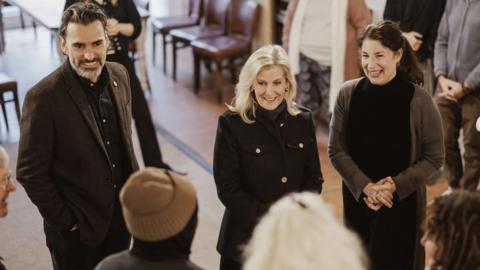 A blonde woman (the Duchess of Edinburgh) in the centre in between a man and a woman talking to other people inside a building.