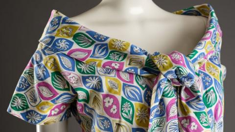 Close-up of the shoulders of a cream mannequin against a dark grey background, wearing a cotton bardot-necked dress with a big tie at the front. The pattern has leaf shapes all over in blue, green, yellow and pink, and within the leaves are other flowers and leaves in white