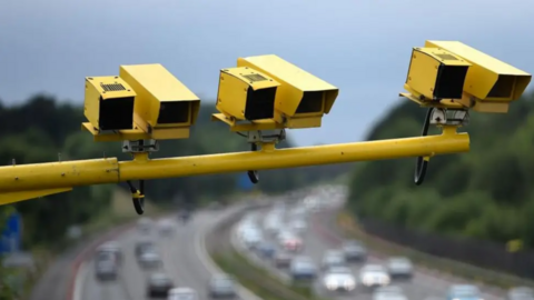 A close-up of yellow average speed cameras which are overlooking a motorway. Blurred motorway traffic can be seen in the background.