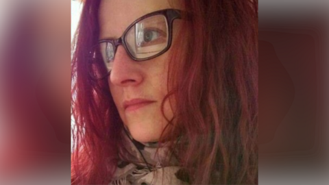 Kelly Anderson is pictured in selfie-style image. She looks away from the camera. She has short red hair, brown glasses and a scarf around her neck.