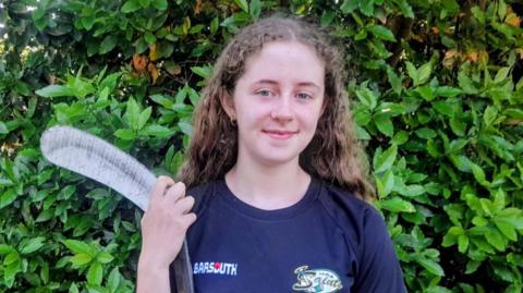 Molly has long brown curly hair and is holding her ice hockey stick.
She is standing outside and is wearing a navy T-shirt.